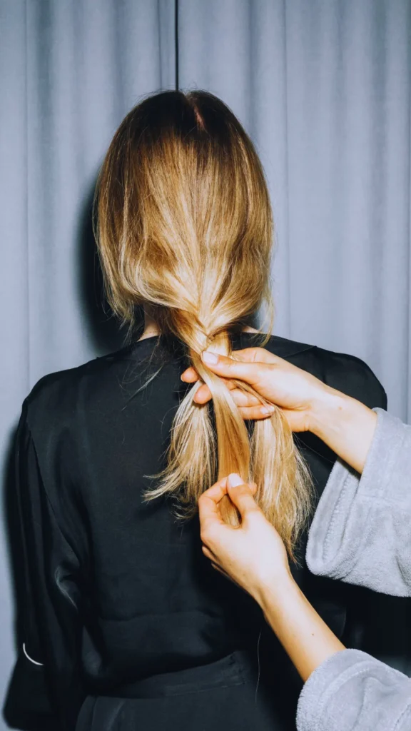 Close-up from behind of a person with long blonde hair being styled into a loose three-strand braid. One hand holds the crossed sections near the nape of the neck while another hand separates a section below, demonstrating the technique against a dark black top and gray curtain background.