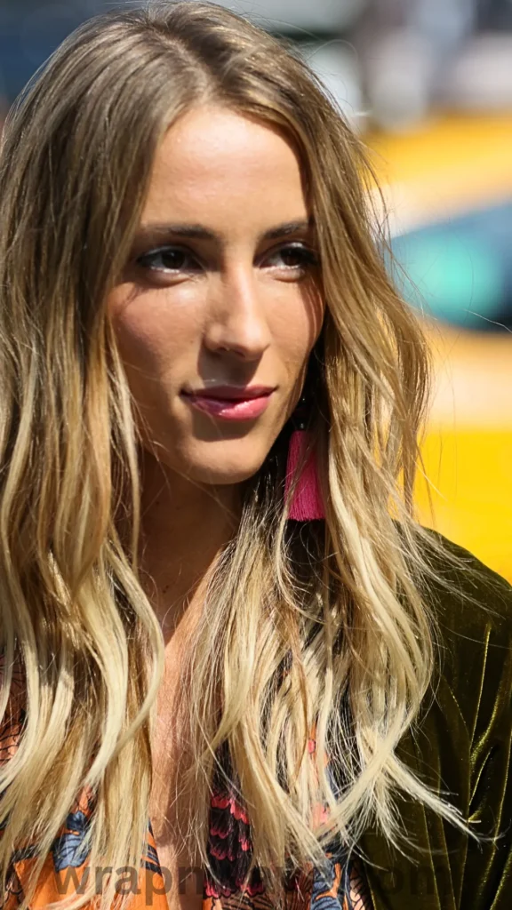 A close-up portrait of a woman looking slightly to the side, showcasing her long blonde hair styled in soft, undone canyon waves with multi-tonal highlights, as referenced in the file 1 (14).webp. She is wearing pink tassel statement earrings and a dark olive-green velvet top over a patterned garment. The shot is captured outdoors in bright sunlight, with a warm, out-of-focus yellow background. A faint "wrapnwave.com" watermark is visible along the bottom edge.