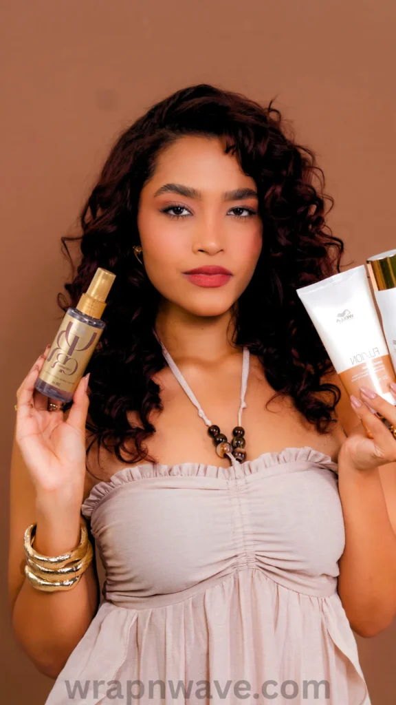 A portrait of a woman with voluminous, dark reddish-brown curly hair posing against a solid light brown background. Looking directly at the camera with a neutral expression, she holds up a gold hair oil bottle in her right hand and a white squeeze tube product in her left hand. She wears a beige strapless, ruched top, chunky gold bracelets, and a beaded necklace. A translucent "wrapnwave.com" watermark is visible along the bottom edge of the image.
