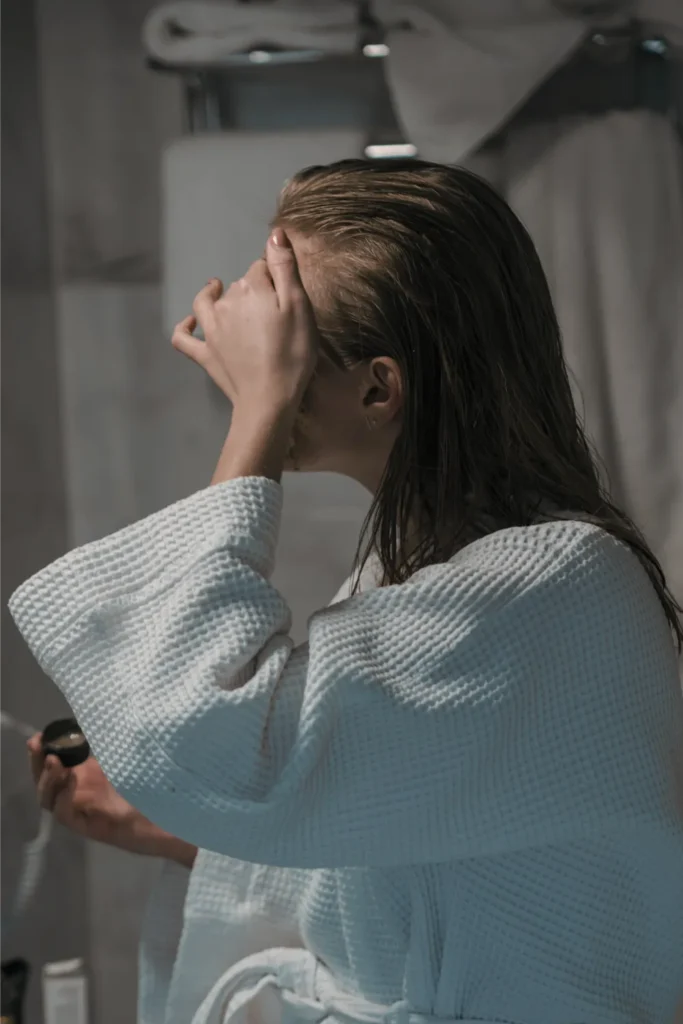 A side profile view of a woman in a white waffle-weave bathrobe standing in a bathroom, applying a hair care product to her long, damp hair before styling. She is holding a small product container in one hand while using the other to distribute the product near her hairline.