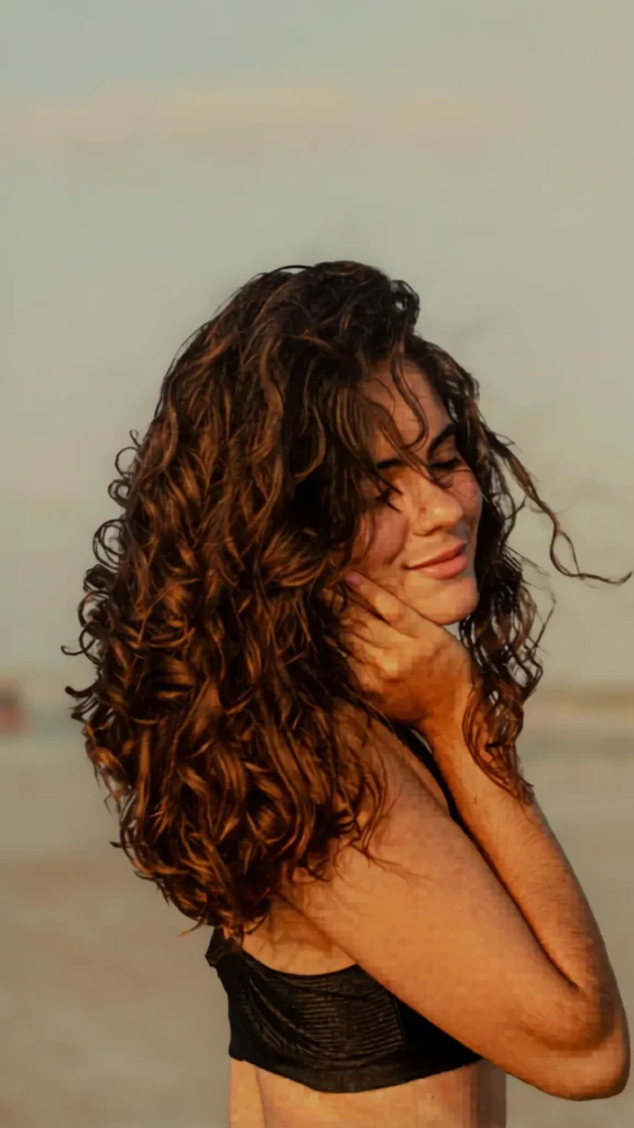 A side profile of a smiling woman with voluminous, highly defined brown curly hair looking down gently. She is touching her face, showcasing frizz-free, shiny, bouncy curls in warm, soft outdoor lighting.