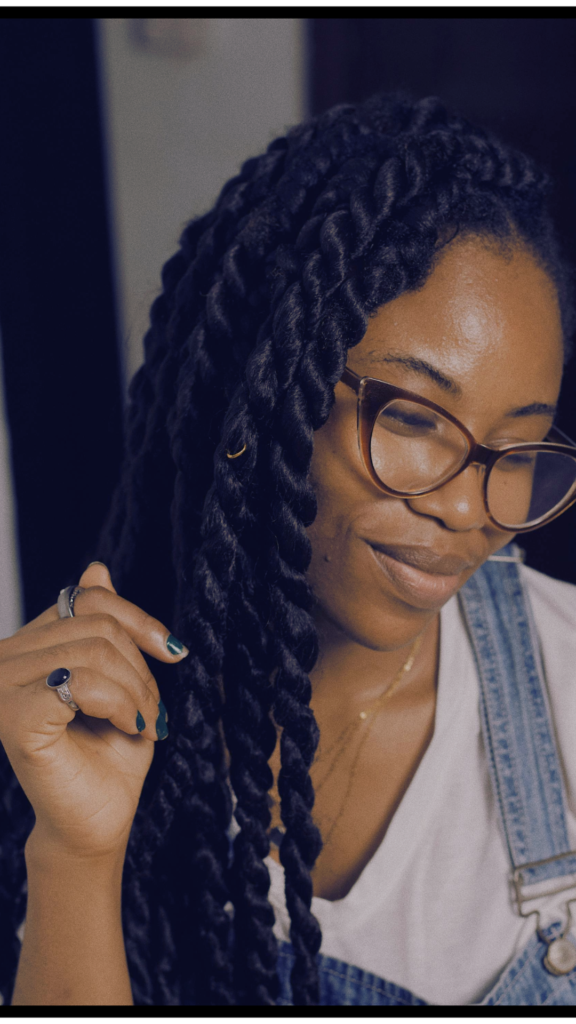 Close-up of a smiling Black woman with long, thick, black twisted braids looking down slightly. She is wearing cat-eye glasses, a white t-shirt, and denim overalls, lightly touching her protective hairstyle which showcases beautiful texture.