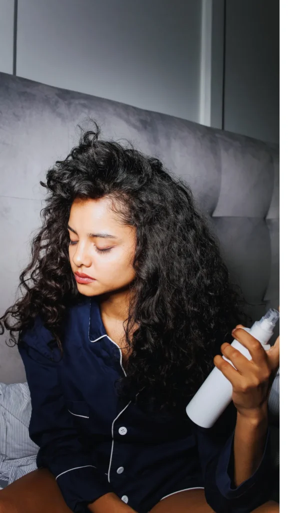 A woman with voluminous, dark curly hair sits on a bed wearing navy blue pajamas with white piping. She is looking down and to the side while holding a white spray bottle in her hand, appearing to get ready for an overnight hair routine.
