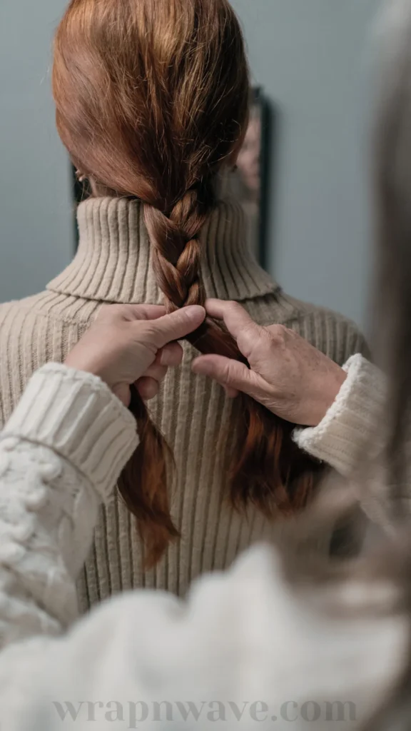 A close-up back view of a woman with long, vibrant red hair wearing a beige ribbed turtleneck sweater. Another person's hands, clad in a white cable-knit sweater, are actively braiding her red hair into a classic three-strand braid down her back. A mirror reflection is subtly visible in the background against a muted grey wall. The text "wrapnwave.com" is watermarked in grey at the bottom, illustrating the process of prepping hair with braids for overnight waves.