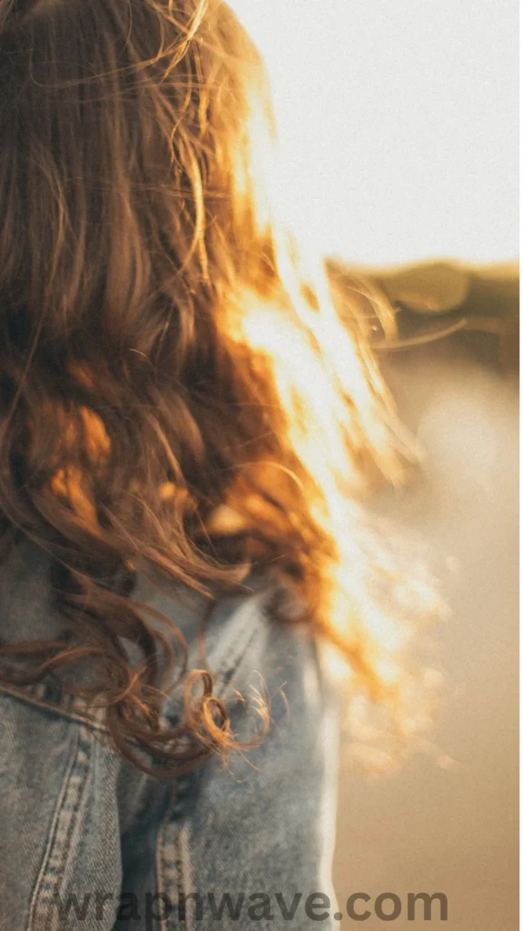 A close-up rear view of a woman with long, voluminous wavy brown hair that is beautifully backlit by warm, glowing golden hour sunlight, highlighting a soft, beachy texture. She is wearing a classic blue denim jacket. The website watermark "wrapnwave.com" is visible at the bottom of the image.