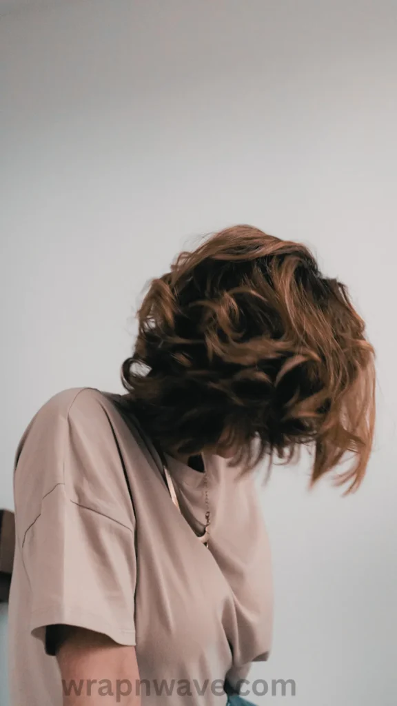A vertical, minimalist photograph focusing on a person with short, voluminous brown hair styled in bouncy, textured heatless curls or waves. The person is sitting and looking downward, completely concealing their face as their loose curls fall forward. They are wearing an oversized, light beige or taupe t-shirt and a simple silver chain necklace against a clean, solid light gray background. A dark "wrapnwave.com" watermark is visible along the bottom edge of the image.