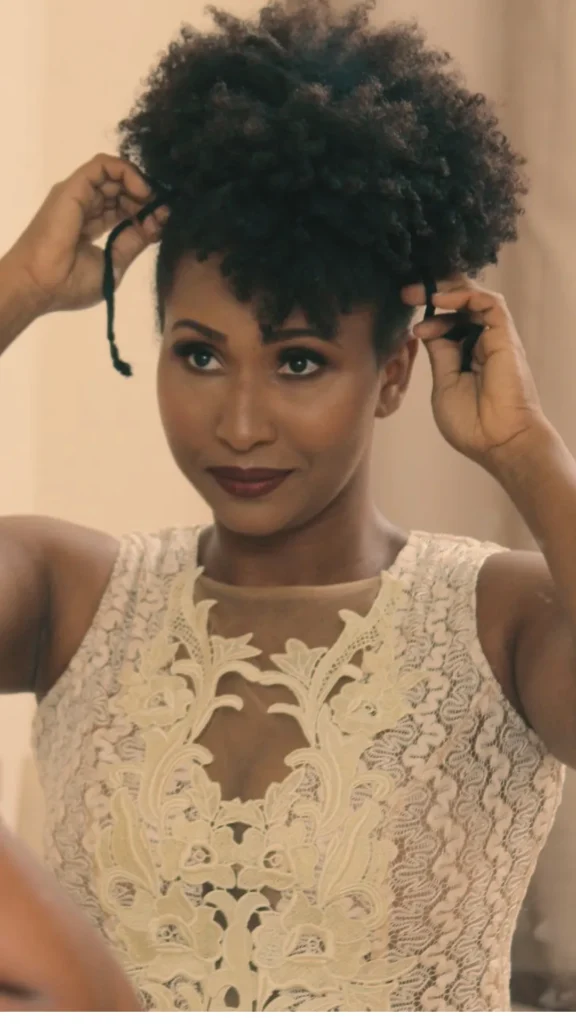 A Black woman with curly natural hair securing a high pineapple hairstyle using a hair tie, demonstrating the original pineapple method popular with curlies.