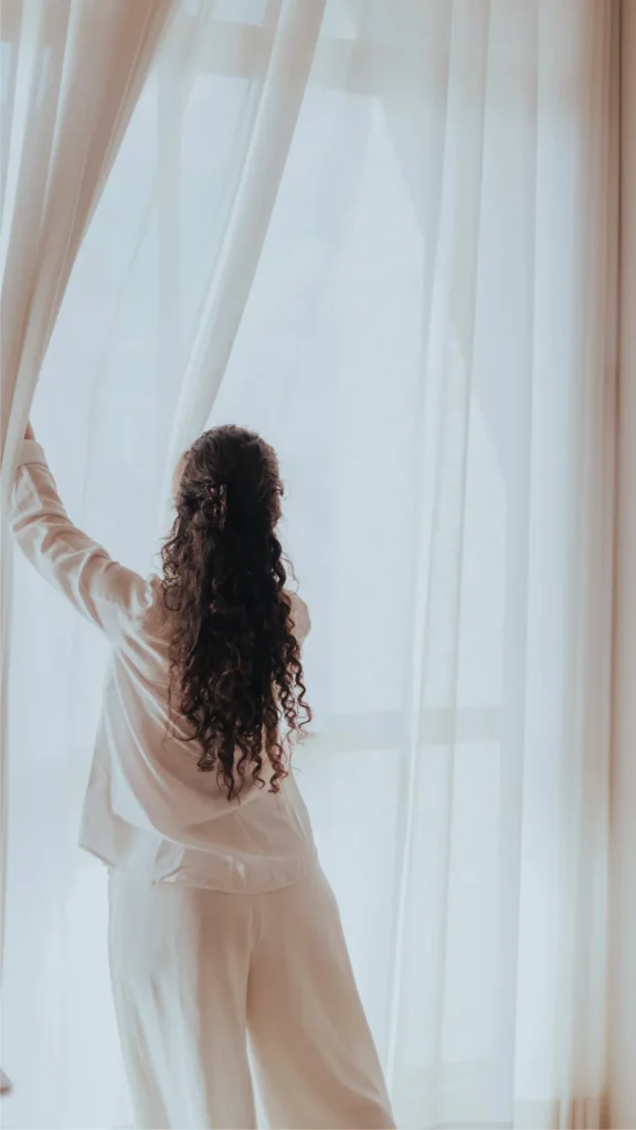 Rear view of a woman with long, voluminous brunette curly hair parted in a half-up style with a small clip. She is wearing comfortable white pajamas and gently parting sheer white curtains to look out a bright window in the morning, showcasing her bouncy, defined overnight waves.