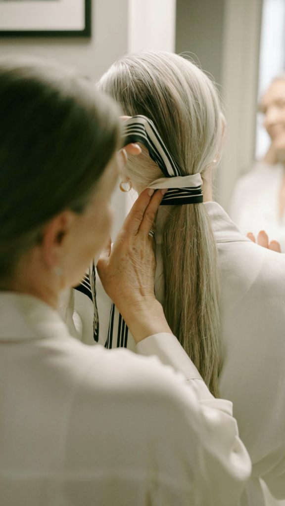 View from behind of a woman with long, straight grey hair having a black-and-white striped fabric scarf wrapped around the base of her ponytail, demonstrating a heatless hair styling technique.