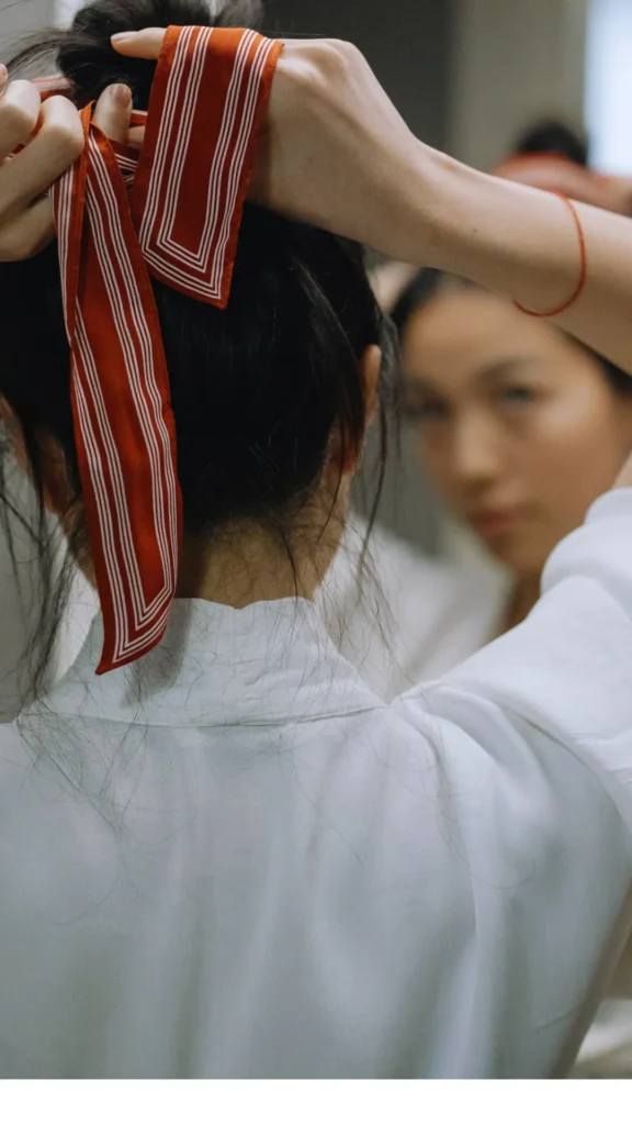 Close-up from behind a woman wearing a white robe as she places a red and white striped silk ribbon or scarf over the top of her dark hair. Her reflection is softly blurred in the mirror ahead, showing her looking at her hands to secure the ribbon for a heatless overnight hair-wrapping routine.