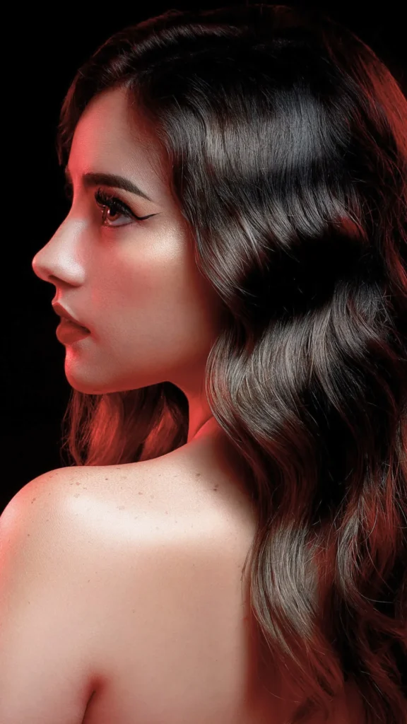 A dramatic profile view of a young woman looking forward, highlighting her long, deep brunette hair styled in glossy, structured waves. Her bare shoulder is slightly turned toward the camera, and the side of her face and hair are illuminated by a moody red rim light against a solid black background.