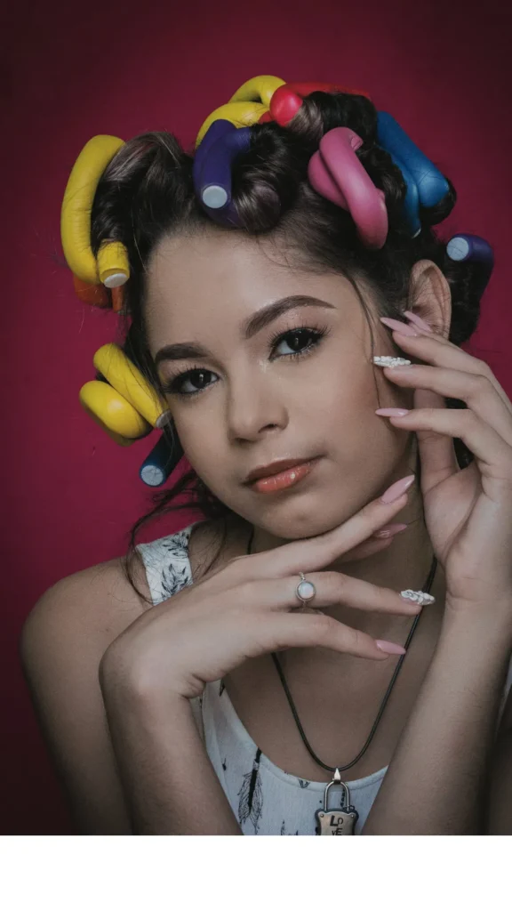 > A portrait of a young woman posing against a vibrant magenta background with her hair wrapped in several bright, multi-colored flexi-rod hair rollers (yellow, purple, pink, and blue). She has her hands framed near her face, showing off long pink nails with white accents, a silver ring, and a lock pendant necklace.