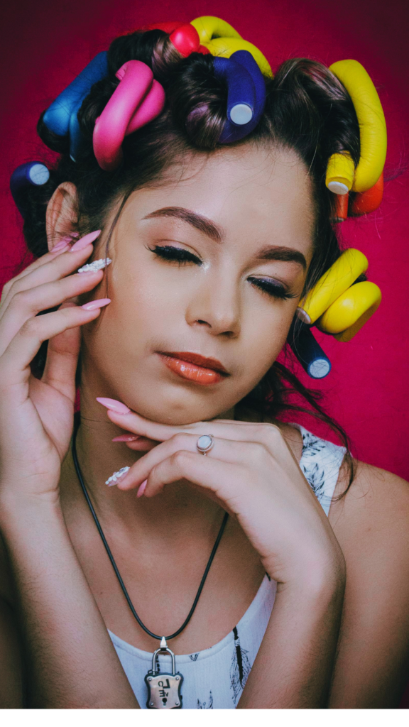 Close-up portrait of a young woman with her eyes closed, showcasing her hair wrapped in vibrant, multi-colored flexi rods (foam rollers) in pink, yellow, and blue for a heatless curling routine.