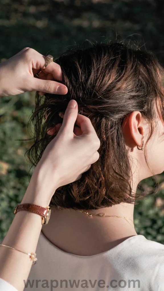A vertical, close-up photograph taken from a side-rear angle, showing a person with short, damp brown hair styled with natural, piecey beach waves. Another person's hands are visible in the frame, gently styling and twisting sections of the hair near the crown. The person styling wears a brown leather watch, a gold bracelet, and a gold ring. The person being styled wears a simple white top, a thin gold necklace, and a small hoop earring. The background features a softly blurred texture of green foliage, and a dark "wrapnwave.com" watermark is visible along the bottom edge of the image.