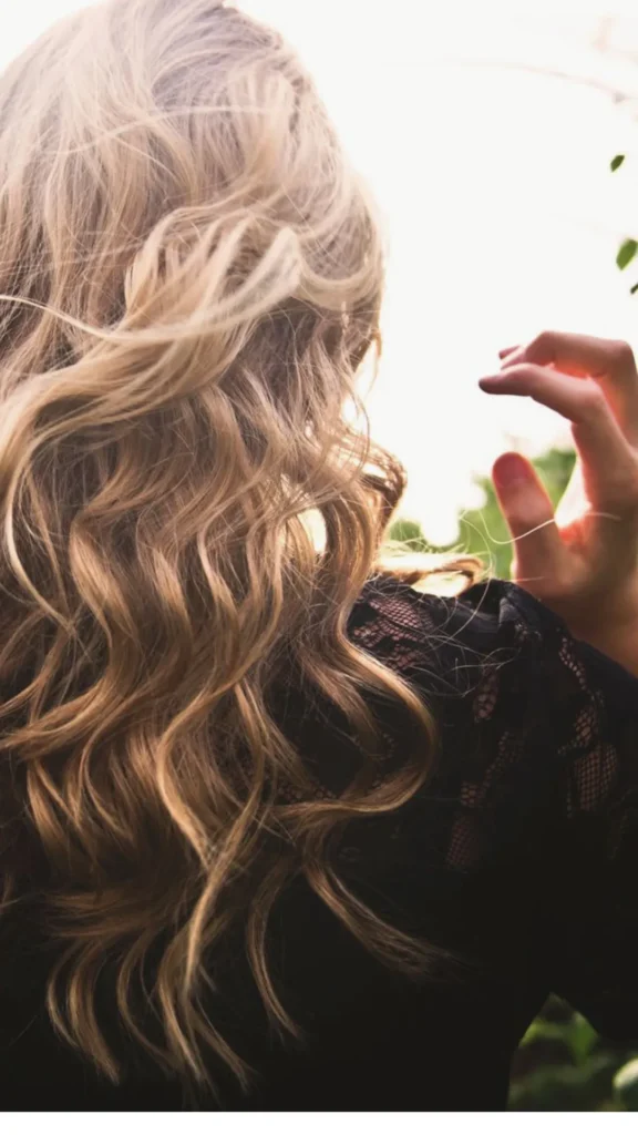 An over-the-shoulder view of a woman with long blonde hair styled into soft, gorgeous waves. She is wearing a black lace top outdoors, with the gentle light illuminating the defined, frizz-free texture of her wavy hair.