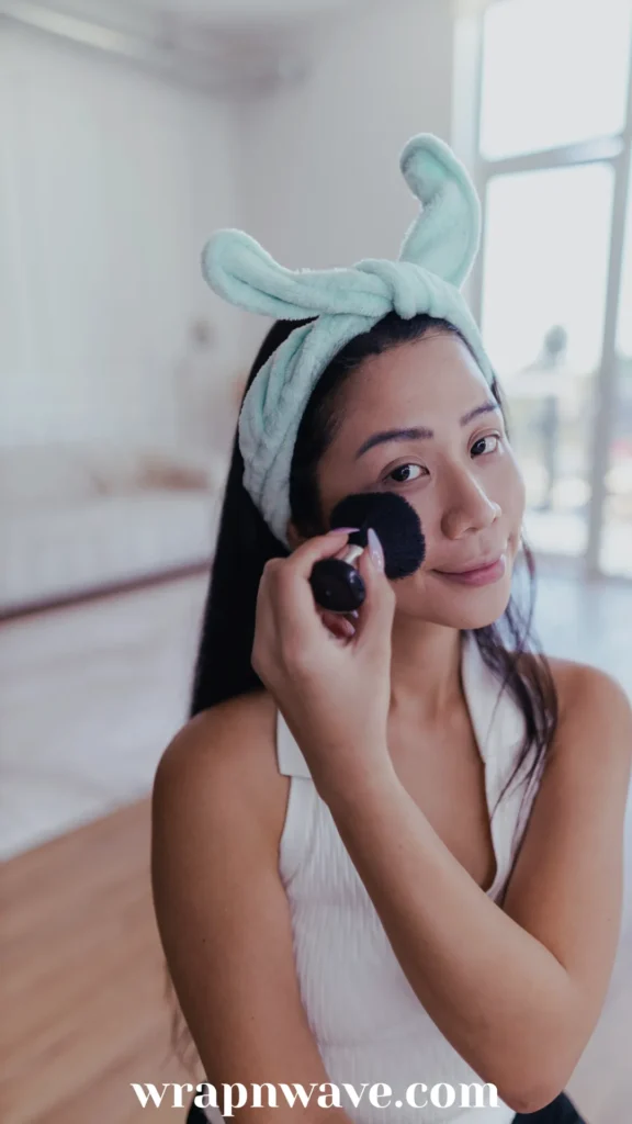 A woman with long dark hair wears a plush, mint green spa headband with a large bow while applying makeup to her cheek with a black powder brush.