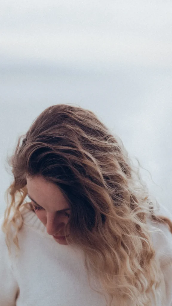A high-angle, close-up shot of a person looking down, highlighting their long, voluminous blonde and light brown hair styled in soft, natural wavy curls. The hair has subtle honey highlights and texture, showcasing the final result of an overnight heatless waves routine. The person is wearing a white knitted sweater against a soft, bright, minimalistic background