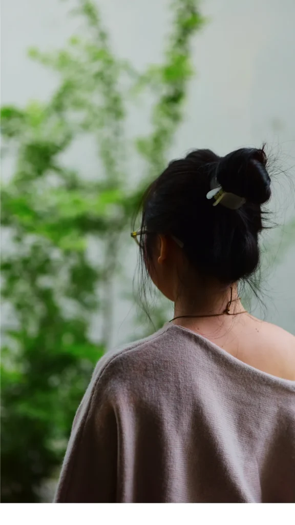 View from behind of a woman with her dark hair twisted into a high, loose bun on top of her head, secured with a small claw clip. She is wearing a cozy, off-the-shoulder beige sweater and facing toward a window with lush green plants blurred in the background.