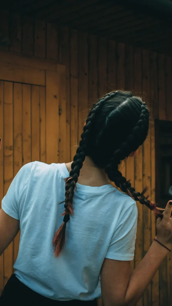 A view from behind of a person with dark hair styled into two tight, symmetrical French braids, also known as boxer braids, starting from the crown of the head. She wears a plain white t-shirt and stands in front of a rustic, textured wooden paneled wall, holding the end of one b