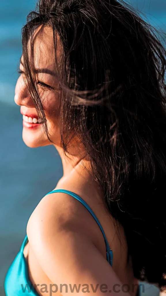 A close-up profile portrait of a smiling woman with long, dark, textured wavy hair blowing across her face in the wind, as referenced in the file 4 (11).webp. She is wearing a bright turquoise or teal bikini top with thin straps, captured in warm, natural sunlight. The background shows a soft-focus blue ocean. A translucent "wrapnwave.com" watermark is visible along the bottom edge.