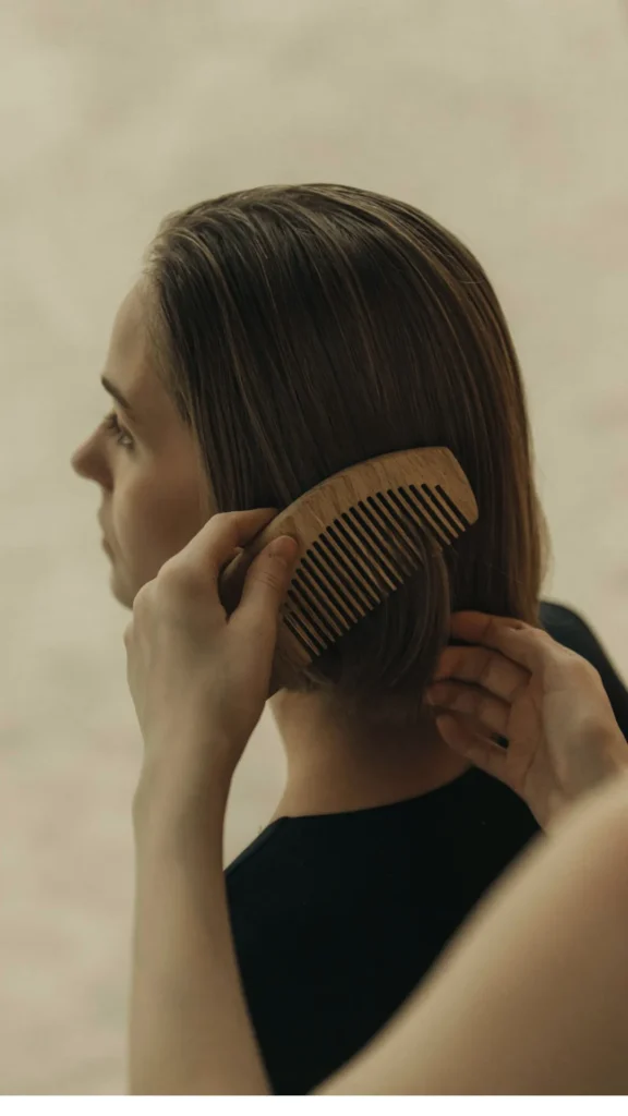 A side profile view of a woman with straight, short brown hair having her hair gently detangled and styled. One hand holds a wide-tooth wooden comb against her hair while another hand guides the strands, illustrating a gentle morning hair care routine.