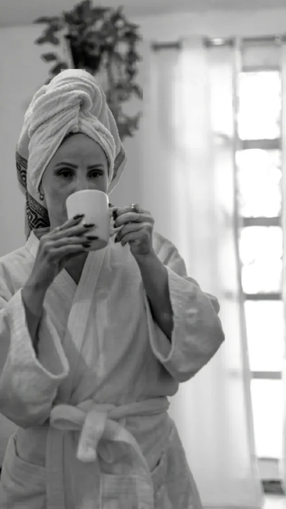 Black and white photo of a woman in a bathrobe with her hair wrapped in a towel, drinking from a mug, setting a morning routine mood for overnight curling methods.