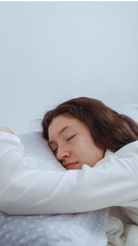 Close-up profile view of a young woman with long, dark brown wavy hair sleeping peacefully on her side. Her head rests on a soft grey and white patterned pillow, and she is wearing a white long-sleeved shirt or pajamas. The minimalist, bright setting emphasizes a calm, friction-free sleeping environment perfect for protecting an overnight hair routine.
