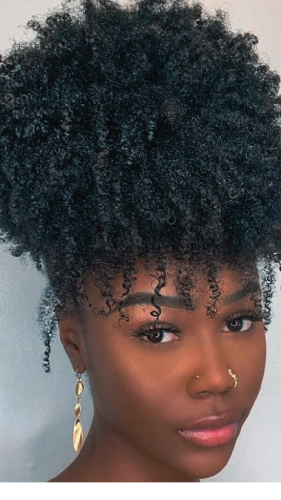 Close-up portrait of a Black woman with natural, highly textured curly hair styled up on top of her head in a voluminous "pineapple" hairstyle, showcasing defined coils and curls forming curly bangs on her forehead.