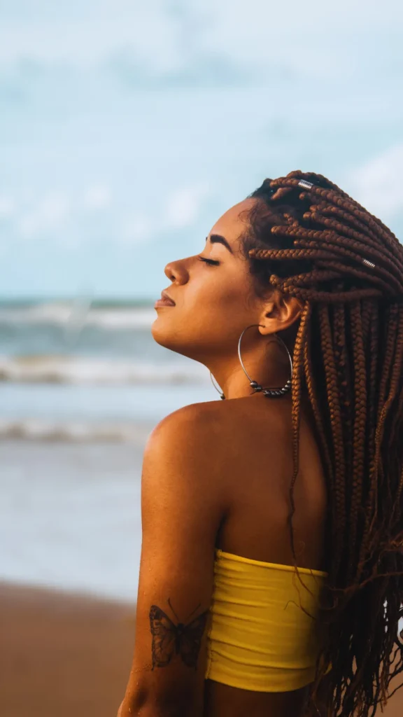 Side profile of a woman with long, golden-brown box braids facing the ocean at the beach with her eyes closed, enjoying the breeze. She is wearing a yellow tube top and large hoop earrings, with a butterfly tattoo visible on her upper arm.