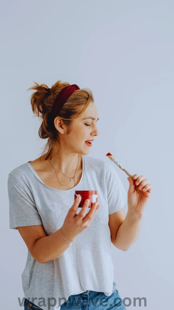 Side profile view of a woman with her blonde hair pulled up into a messy bun, secured with a wide, dark red or burgundy fabric headband. She is wearing a grey t-shirt, blue jeans, and delicate silver necklaces against a plain white background. She is holding a small red jar in one hand and an artist's paintbrush with red paint on the bristles in the other, speaking or looking at the brush. The website watermark "wrapnwave.com" is visible at the bottom.