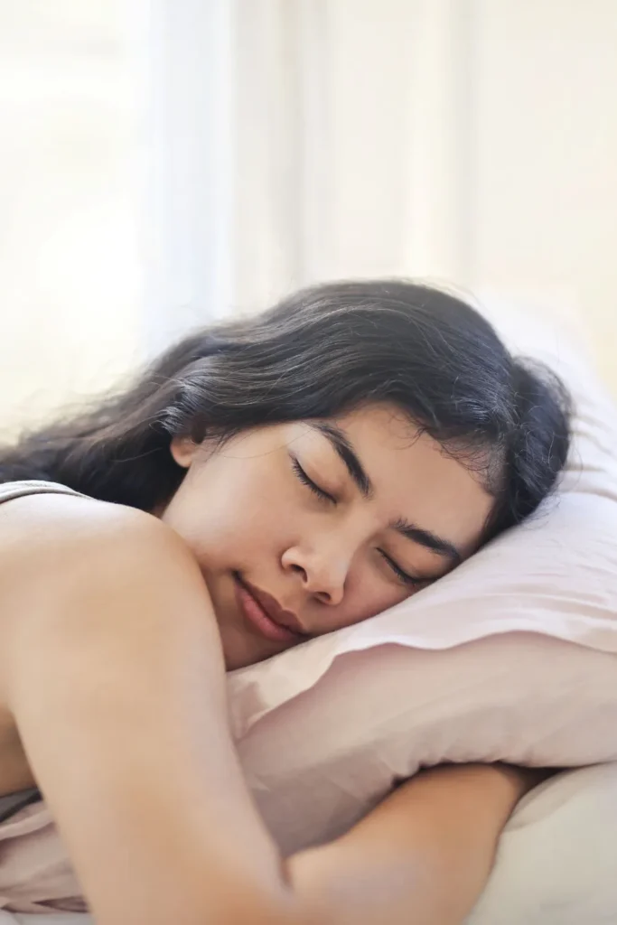 A close-up view of a peaceful woman with dark hair sleeping comfortably on her side. Her head is resting on a soft, light pink satin or silk pillowcase, illustrating a frictionless and hair-protective nighttime sleep routine.