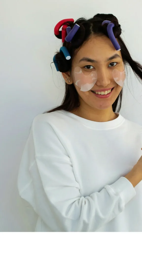 A smiling East Asian woman in a white sweatshirt wearing colorful flexi rods in her hair and under-eye patches, demonstrating long hair styling tips for overnight beach waves.