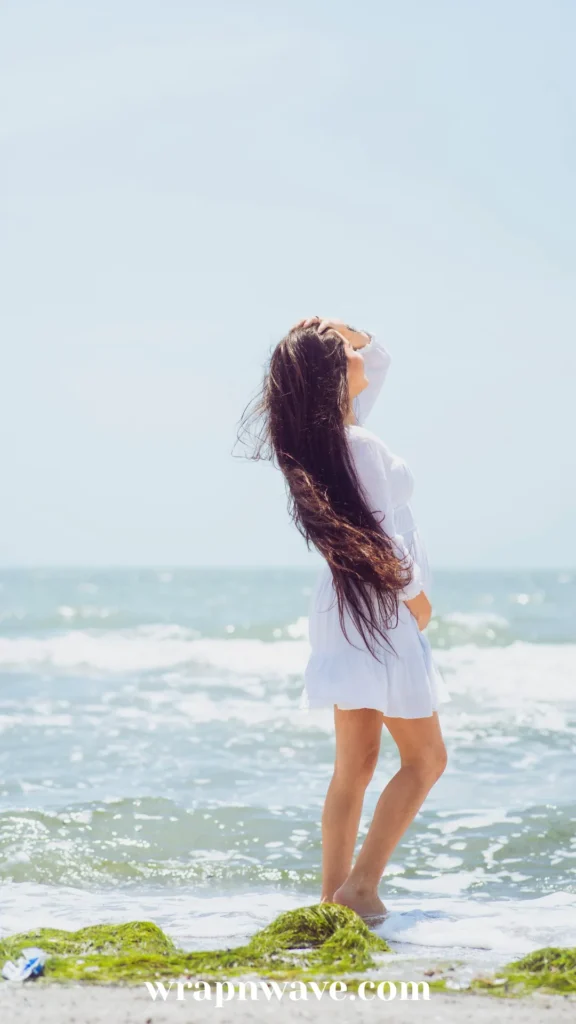 Side profile of a woman with long, flowing dark brown hair standing on a beach shore wearing a short white dress, running her hand through her hair with ocean waves in the background.