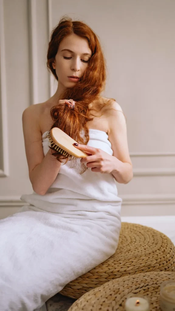A woman with long, vibrant red hair wrapped in a white bath towel sits on a woven straw pouf ottoman. She looks down as she uses a wooden paddle hairbrush to gently brush out the ends of her wavy hair, with a small candle glowing softly on the floor next to her.