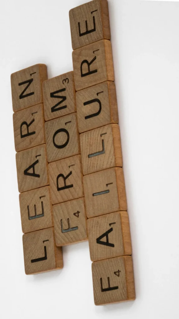A top-down, slightly angled view of wooden letter tiles arranged on a plain white background to spell out the phrase "LEARN FROM FAILURE". The tiles resemble Scrabble pieces with small numbers on them. The image serves as a conceptual visual for troubleshooting mistakes and learning how to fix overnight hair routine mishaps.