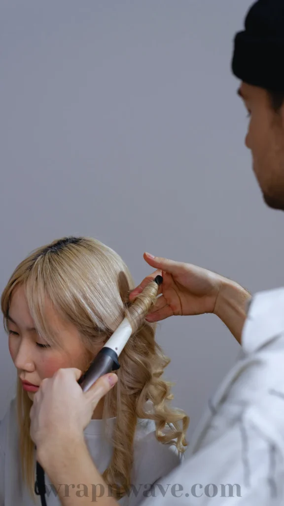 A medium close-up of a young East Asian woman with blonde hair sitting down with her eyes lowered. From a side-profile view, a stylist wearing a black beanie and white shirt is shown carefully wrapping a section of her hair around a heated curling wand to create defined curls. The background is a solid, neutral grey. The text "wrapnwave.com" is watermarked in grey at the bottom, illustrating a traditional heated styling method for creating waves.