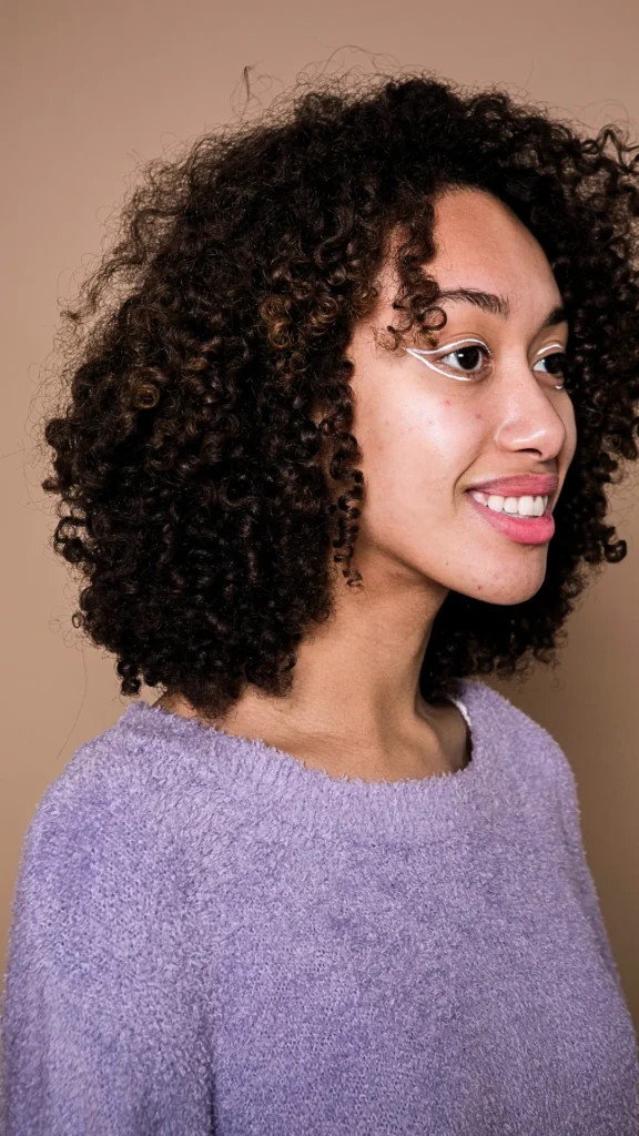 A three-quarter profile studio portrait of a smiling young woman with voluminous, highly defined natural curls and coils. Her dark brown hair features subtle caramel highlights and is styled into a rounded curly shape. She is wearing a fuzzy purple crew-neck sweater and unique white graphic eyeliner accents under her eyes, set against a solid beige backdrop.