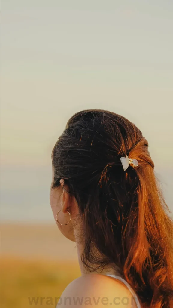 A vertical photograph taken from behind a woman with long brown hair, as referenced in the file 5 Easy Ways to Get Beach Waves on a Bob-Zero Heat Damage! (7).webp. Her hair is loosely gathered into a half-up, half-down style, secured neatly at the back of her head with a small, light-colored butterfly claw clip. She wears thin gold hoop earrings and looks out toward a soft, hazy horizon during a warm golden hour or sunset. A faint, semi-transparent "wrapnwave.com" watermark is visible along the bottom edge of the image.