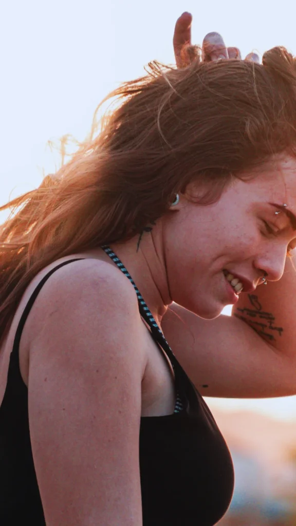 A side profile shot of a smiling young woman with long, textured brown hair, illuminated by warm golden hour sunlight. She has her hand running through her hair at the crown, effortlessly shaking out and reviving her natural, windswept waves. She is wearing a black tank top and has visible tattoos and facial piercings, creating a casual, carefree morning vibe.