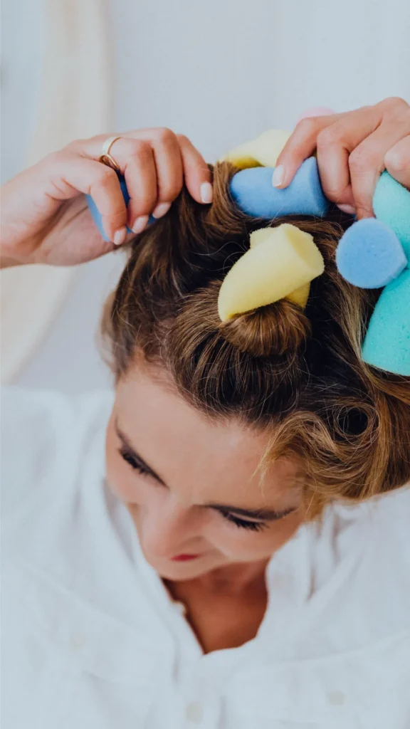 A top-down view of a woman with light brown hair rolling sections of her hair into colorful foam curlers, demonstrating a heatless method for frizz-free beach waves.