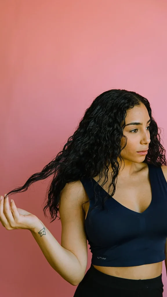 A portrait of a woman with long, textured, dark wavy hair looking slightly to the side against a solid pink background. She is holding a single strand of her wavy hair with her hand, showing a small line tattoo of an animal face on her wrist. She is wearing a dark blue athletic V-neck tank top and black pants.