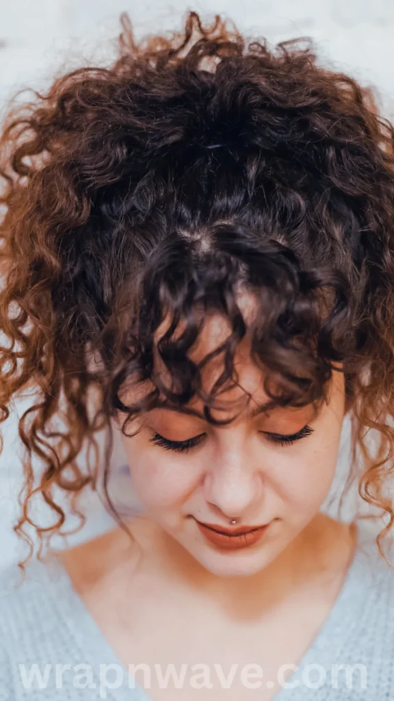 A close-up, high-angle portrait of a young woman with voluminous, highly defined dark curly hair pulled up into a high pineapple ponytail or half-up style, as referenced in the file wrap & (26).webp. She is looking downward with her eyes closed, showing her long lashes, warm eyeshadow, and a small dermal piercing right below her bottom lip. Several tight curls fall forward onto her forehead as bangs, and a translucent "wrapnwave.com" watermark is visible along the bottom edge of the image.