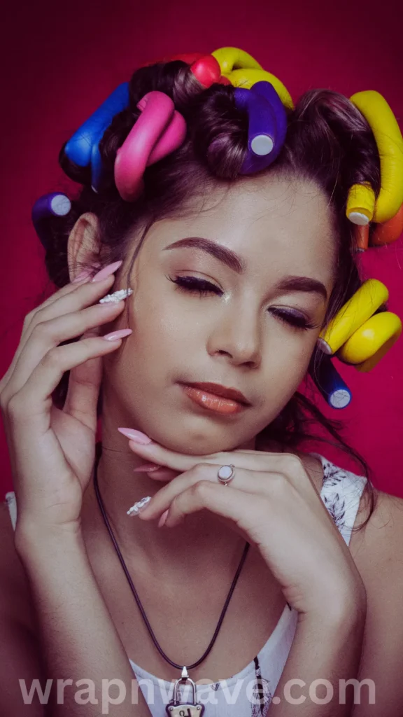 A close-up portrait of a young woman posing with her eyes closed against a vibrant pink background. Her hair is wrapped in numerous bright, multicolored flexible foam curling rods in shades of yellow, pink, blue, purple, and green. She holds both hands up to her face, showcasing long pink manicured nails with white 3D floral accents on her ring fingers. She is wearing a white printed top, a black cord necklace with a lock pendant, and a simple ring on her finger. A translucent "wrapnwave.com" watermark is visible along the bottom edge of the image