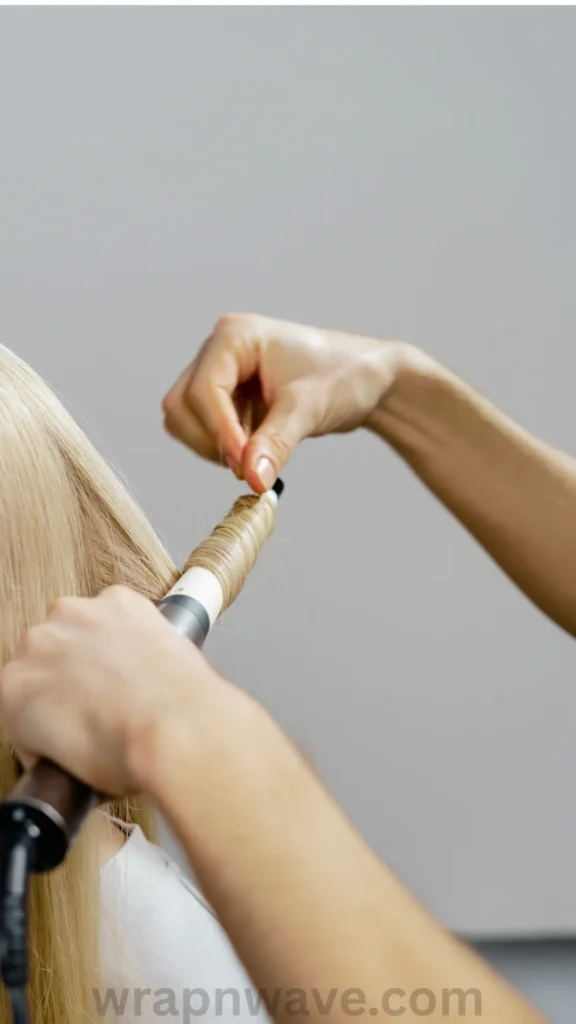 A close-up shot showing a person's hands wrapping a section of straight blonde hair tightly around the barrel of a hot curling iron or wand, as referenced in the file wrapnwave.com (14).webp. The background is a solid, neutral grey wall, and a translucent "wrapnwave.com" watermark is visible along the bottom edge of the image.