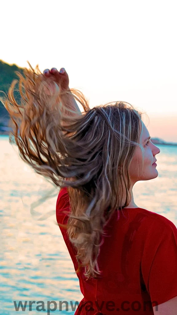 A side-profile photograph of a woman with long, textured blonde and brown highlighted hair, as referenced in the file wrapnwave.com (16).webp. She is wearing a bright red top and stands outdoors by the sea, running her hand through her hair to flip it upward, creating dynamic movement. The background features a soft-focus body of water reflecting the light of a pale, warm sky at sunset or sunrise, and a translucent "wrapnwave.com" watermark is printed across the bottom edge of the image.