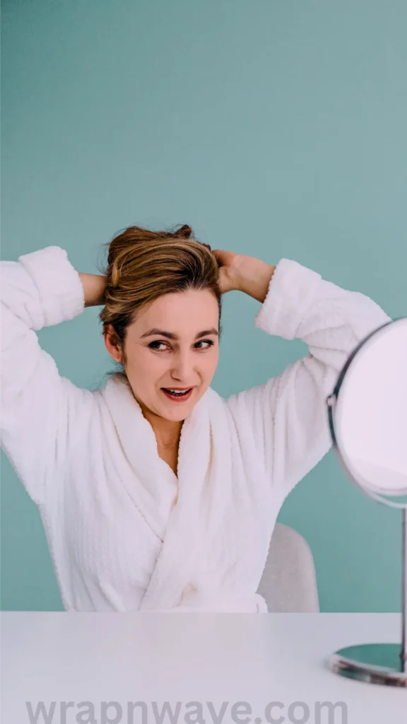 A front-facing photograph of a smiling woman sitting at a white table, as referenced in the file wrapnwave.com (17).webp. She is wearing a soft, plush white bathrobe and has both hands raised behind her head, styling or gathering her light brown highlighted hair. She looks slightly to the right toward a round, silver-framed vanity mirror resting on the table. The entire scene is set against a solid, light teal-blue background wall, and a translucent "wrapnwave.com" watermark is visible along the bottom edge of the image.