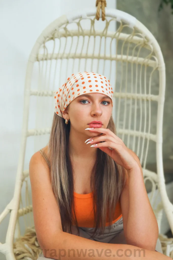 A vertical, close-up photograph of a young woman with long, straight light brown or blonde hair looking forward, as referenced in the file wrapnwave.com (26)_2.webp. She is wearing a cream-colored headscarf decorated with bright orange polka dots tied over her head. Her hand, showcasing long white stiletto nails, rests gently under her chin as she poses in an orange tank top. She is seated in a white woven wicker hanging egg chair. A semi-transparent "wrapnwave.com" watermark is visible along the bottom edge of the image.