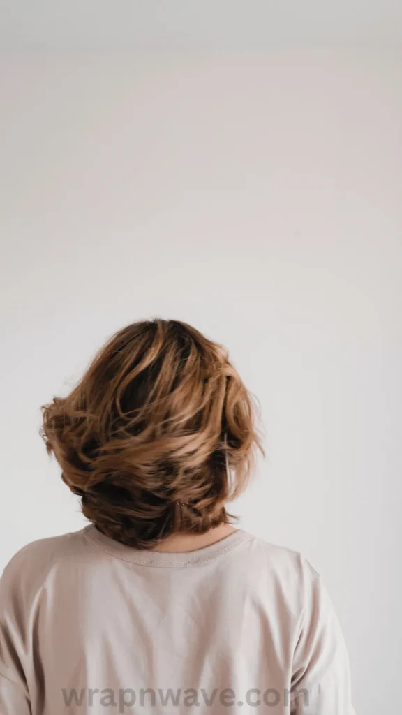 A vertical photograph taken from behind a person showcasing a styled, wavy short haircut, as referenced in the file wrapnwave.com (28) (1).webp. The hair is a dimensional light brown or dark blonde color with soft highlights, styled into loose, piecey layers and voluminous waves that fall just above the shoulders. The person is wearing a casual, light beige t-shirt and posing against a clean, plain white background. A semi-transparent "wrapnwave.com" watermark is visible along the bottom edge of the image.
