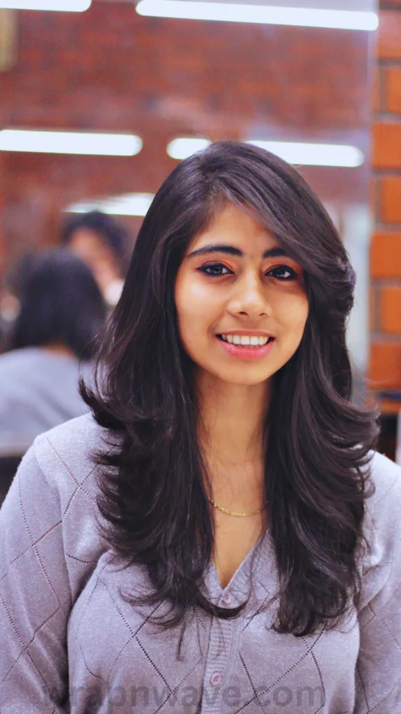 A vertical, chest-up photograph of a smiling young woman with long, voluminous dark hair looking directly at the camera, as referenced in the file wrapnwave’com (1).webp. Her hair is styled with a deep side part and features prominent, outward-curling layers that frame her face and shoulders. She has a bright smile, eye makeup, and is wearing a thin gold necklace paired with a light gray, textured long-sleeved top with a subtle diamond pattern. The background shows a softly blurred indoor environment with exposed brick accents and warm lighting. A semi-transparent "wrapnwave’com" watermark runs along the bottom edge of the image.