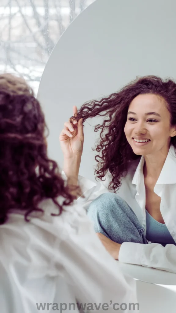 A vertical, close-up photograph taken from behind a person with long, voluminous curly dark brown hair looking into a round mirror, as referenced in the file wrapnwave’com.webp. The reflection shows a smiling young woman wearing an open white collared shirt over a light blue top and denim jeans. She is holding up a section of her tightly defined, bouncy curls with her right hand while admiring them in the mirror. The background is minimal and bright, and a semi-transparent "wrapnwave’com" watermark runs along the bottom edge of the image.